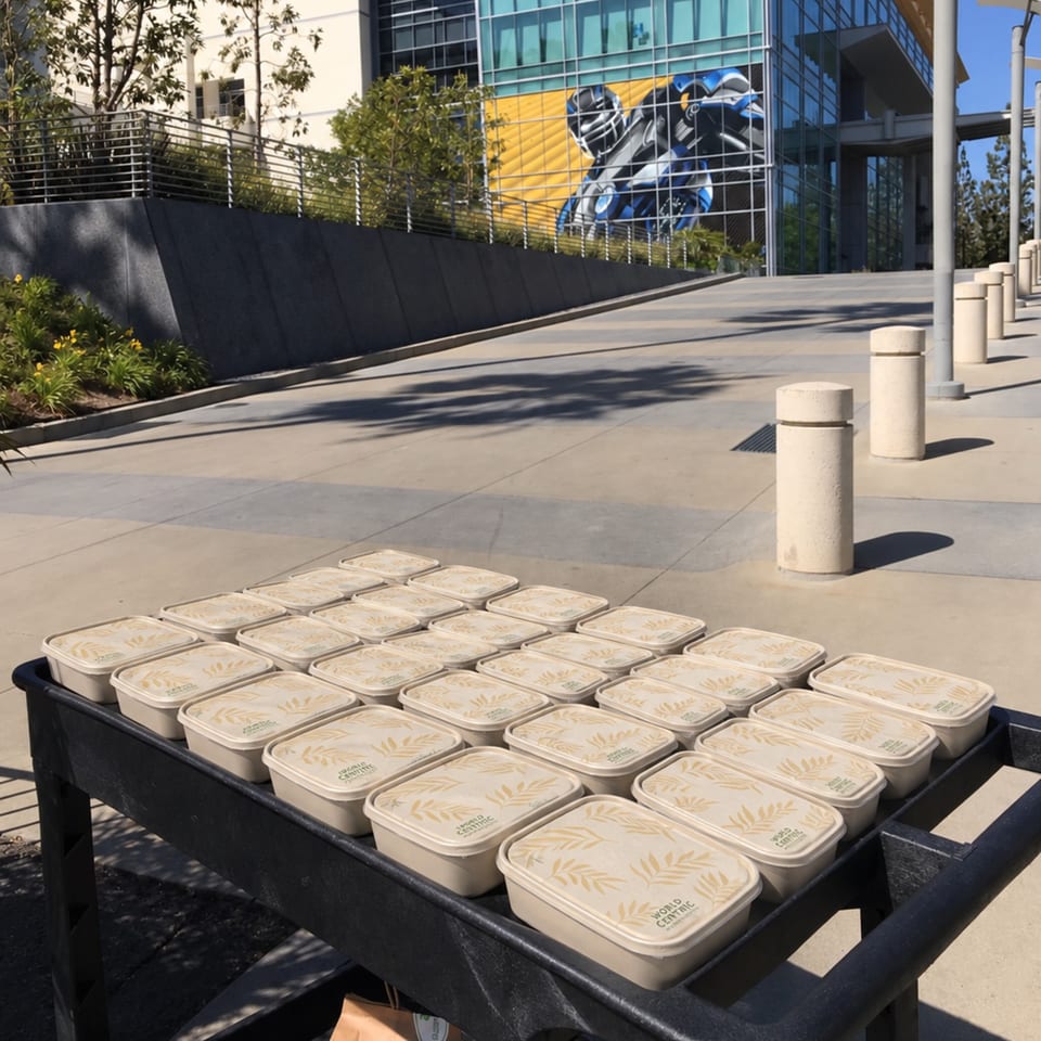 A black cart holds rows of compostable meal containers outside a modern building with a large blue-and-yellow mural.